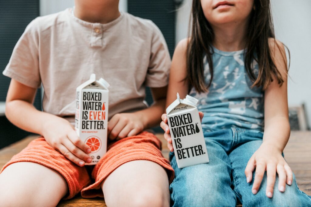 a boy and a girl sitting on a table holding boxes of booze
